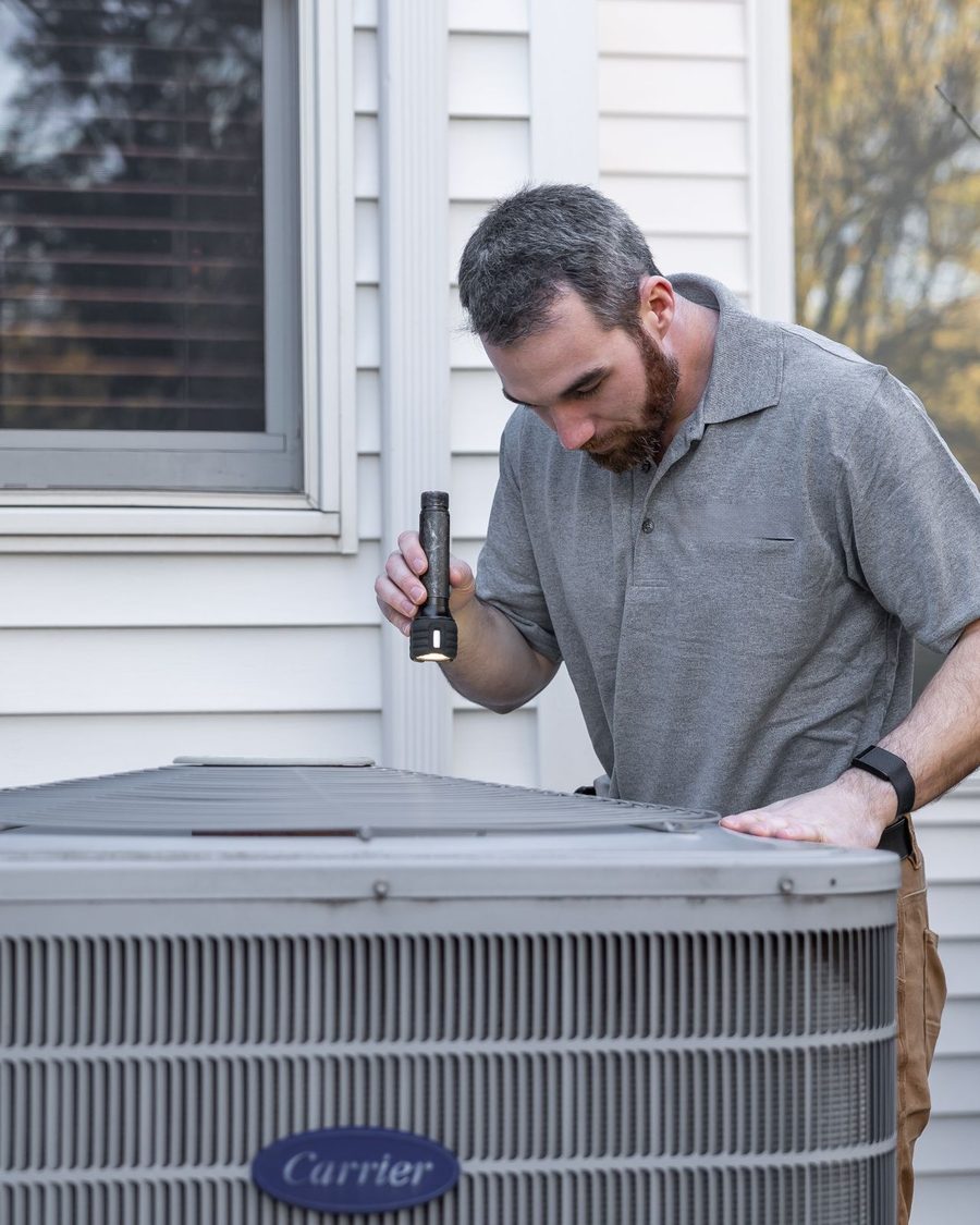 A technician with a flashlight inspecting an outdoor condenser unit beside a house